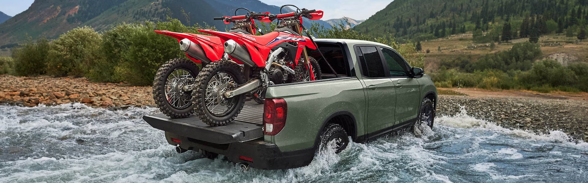 2026 Honda Ridgeline pickup truck carrying dirt bikes through shallow water crossing.